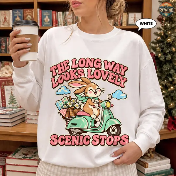 Woman holding a coffee cup wearing a white crewneck sweatshirt printed with a cheerful bunny riding a mint scooter and the words The Long Way Looks Lovely Scenic Stops.