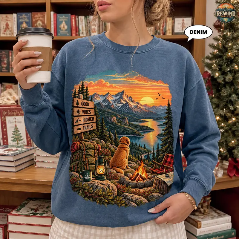 Model wearing a denim blue crewneck sweatshirt with a sunset mountain lake print featuring a dog, campfire, backpack and wooden trail signs.