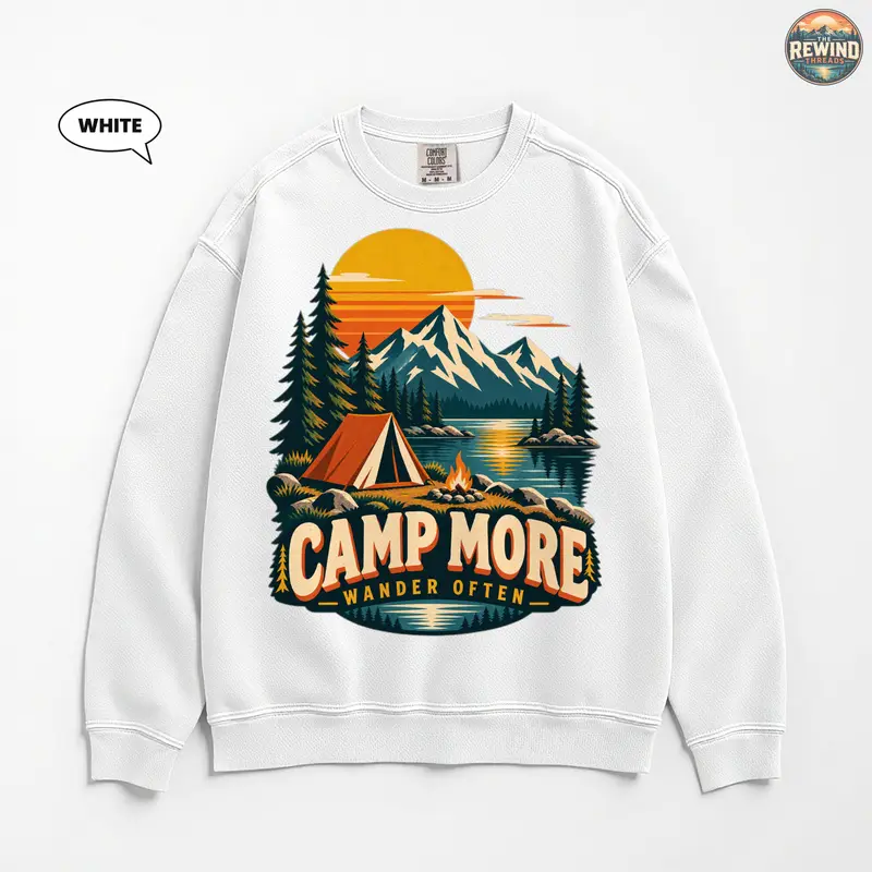 White crewneck sweatshirt laid flat on a white background, featuring a retro mountain lake camping scene with a golden sunset, orange tent and "Camp More Wander Often" lettering.