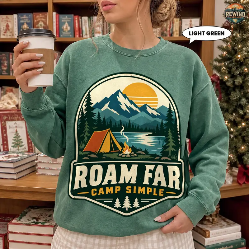 Model wearing a light green crewneck sweatshirt with a vintage camping badge featuring a mountain sunset, forest, lake, orange tent and campfire with "Roam Far, Camp Simple" text.