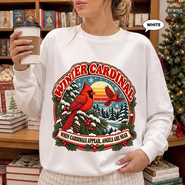 Model wearing a white crewneck sweatshirt with a retro Winter Cardinal graphic featuring two red birds, a snowy evergreen forest, holly and a striped sunset.