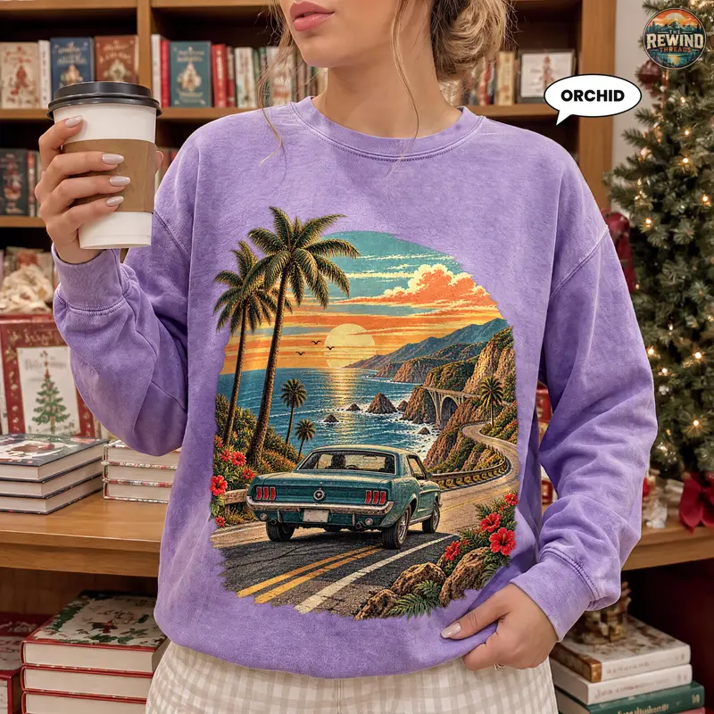 Woman wearing a light purple orchid crewneck sweatshirt with a vintage coastal road scene of a classic car, palm trees and sunset cliffs