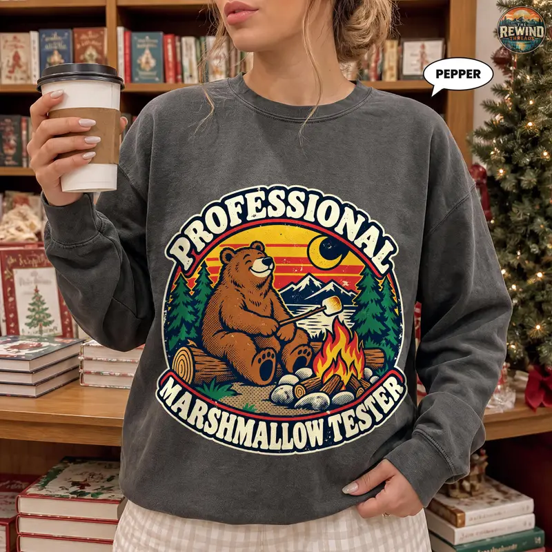 Model wearing a charcoal gray crewneck sweatshirt with a retro bear roasting a marshmallow beside a campfire, mountains and pine trees, with Professional Marshmallow Tester text.