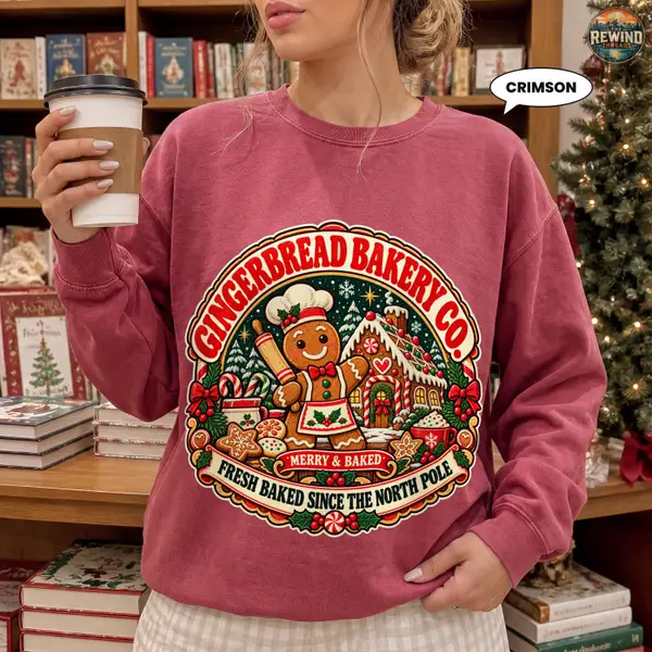 Woman holding a coffee cup wearing a crimson red crewneck sweatshirt with a vintage gingerbread man baking badge print in a cozy holiday setting.