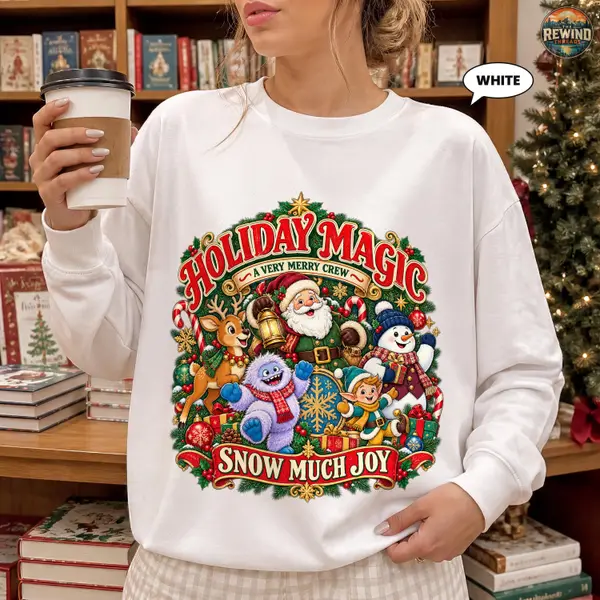 Woman in a white Christmas sweatshirt printed with a Holiday Magic Snow Much Joy design of Santa, reindeer, snowman and snow creatures, holding a coffee cup by a decorated tree.