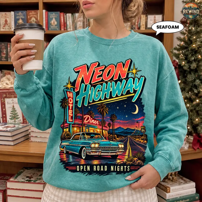 Model wearing a seafoam crewneck sweatshirt with a Neon Highway print featuring a classic car, neon diner and motel signs, palm trees, and a desert road at night.