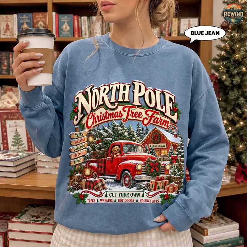 Woman holding coffee wearing a blue jean crewneck sweatshirt printed with a vintage North Pole Christmas Tree Farm badge, red truck, snowy trees and cabin.