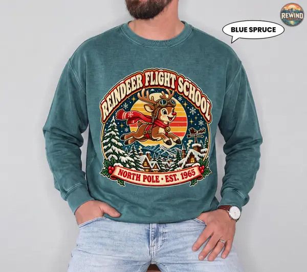 Blue Spruce Christmas crewneck sweatshirt worn by a seated male model with jeans, featuring a retro Reindeer Flight School graphic with a flying reindeer and snowy village.