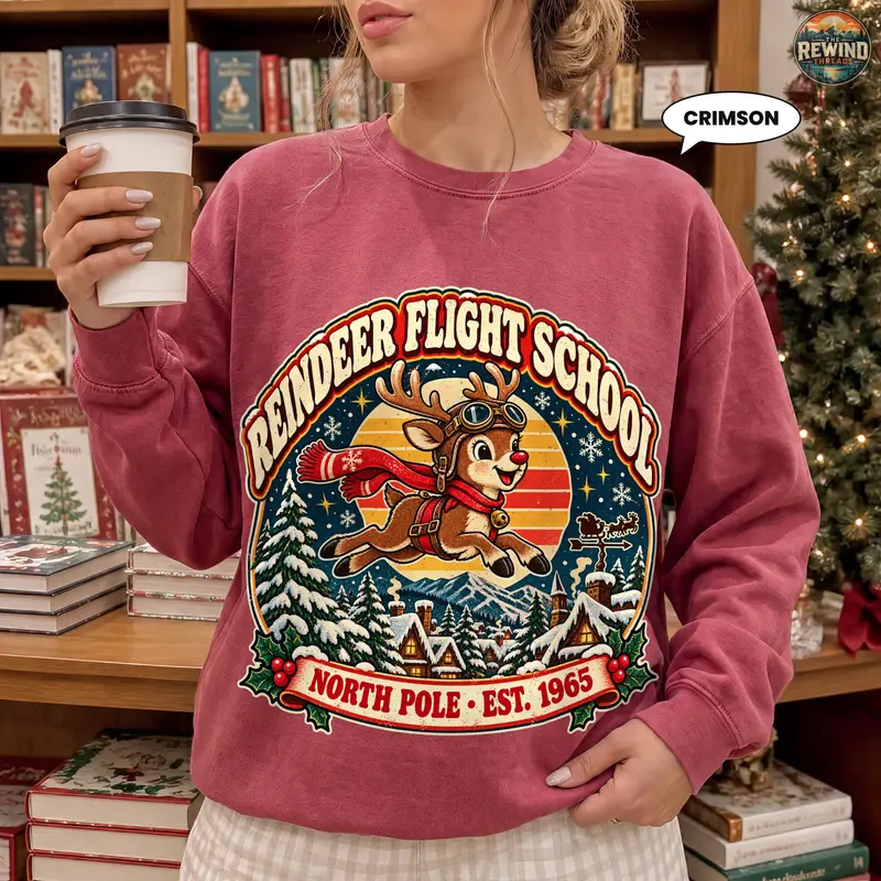 Model wearing a washed crimson crewneck sweatshirt with a retro Reindeer Flight School design featuring a scarf-wearing reindeer, snowy trees and a North Pole banner.