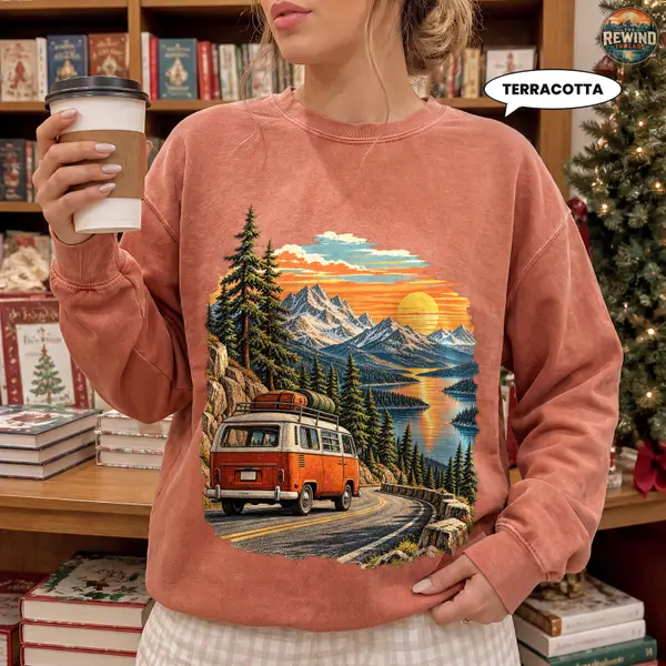 Model wearing a terracotta crewneck sweatshirt with a retro orange camper van print featuring a winding mountain road, pine forest, lake and sunset.