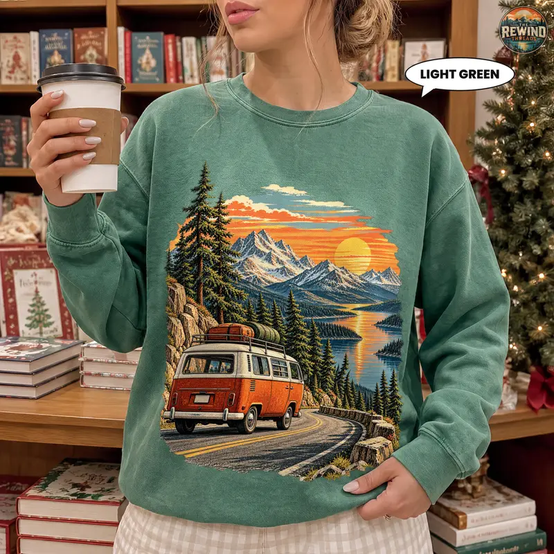 Woman in a light green sweatshirt featuring a retro van on a mountain road with sunset, lake and pine trees