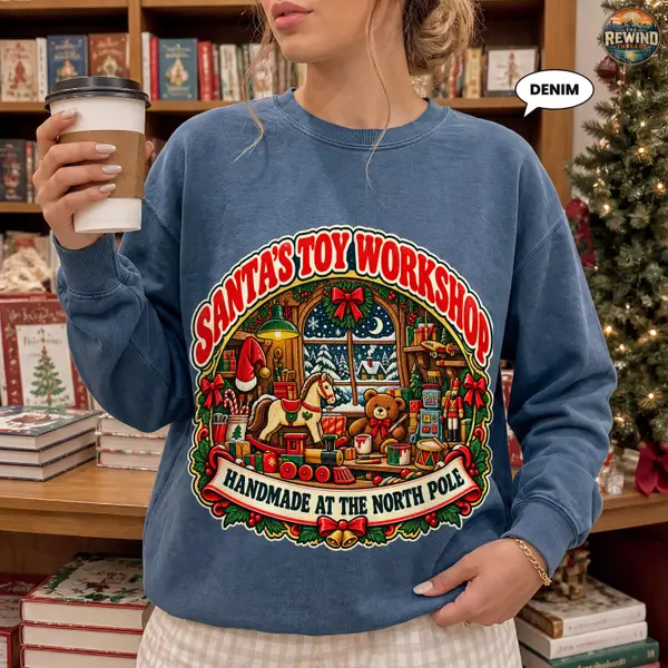 Denim Christmas crewneck sweatshirt worn by a model beside a Christmas tree, featuring a retro Santa's Toy Workshop design with colorful toys, holly and red bows.