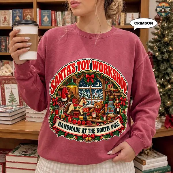 Model wearing a muted crimson crewneck sweatshirt with a retro Santa’s Toy Workshop graphic, colorful toys, holiday bows and a North Pole banner.