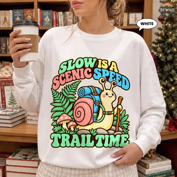 Woman wearing a white crewneck sweatshirt with a retro hiking snail carrying a backpack among ferns and mushrooms and the words Slow Is a Scenic Speed Trail Time