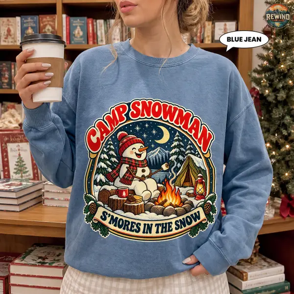 Model wearing a faded blue crewneck sweatshirt with a vintage Camp Snowman print featuring a snowman roasting marshmallows beside a snowy campsite and the words S’mores in the Snow.