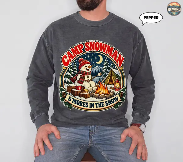 Seated model wearing a pepper charcoal crewneck sweatshirt with a Camp Snowman graphic featuring a snowman, winter campfire, tent and crescent moon.
