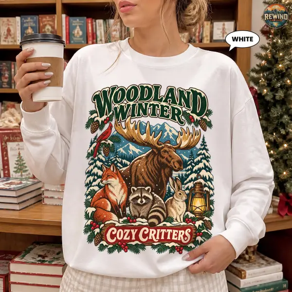 Woman wearing a grey crewneck sweatshirt with a retro snowy forest moose, fox, raccoon and rabbit design and the words Woodland Winter Cozy Critters.